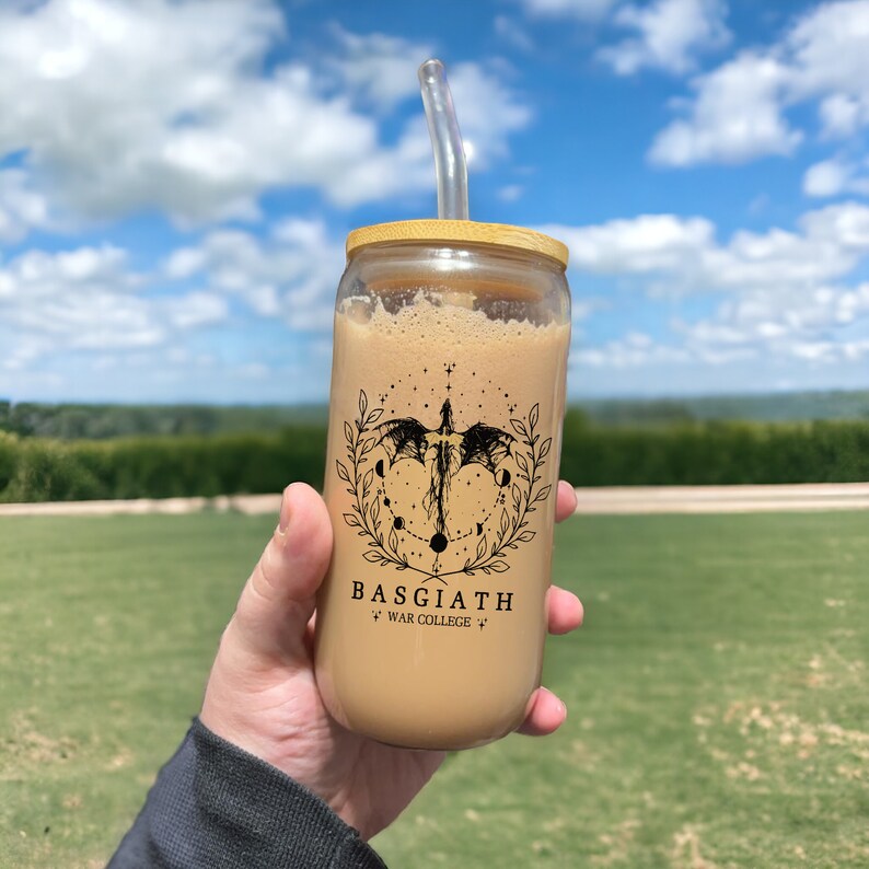 May include: A glass jar with a bamboo lid and a straw, filled with a brown beverage. The jar has a black and white graphic of a dragon with a circle of stars and moons around it. The text "BASGIATH" and "WAR COLLEGE" is printed below the graphic.