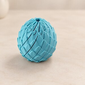 May include: A bright blue, spherical object with a textured, scale-like surface pattern. The item features a series of interlocking, overlapping segments that create a repeating geometric design across its entire rounded exterior.