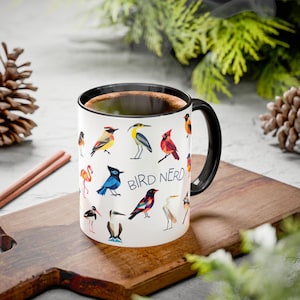 Puede incluir: Taza de cerámica blanca con asa y borde negros, llena de café. La taza está decorada con ilustraciones coloridas de varios pájaros y el texto "BIRD NERD". La taza está sobre una tabla de cortar de madera.