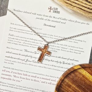 May include: A silver chain necklace with a cross pendant. The cross is brown with a gold center and is set against a printed devotional. The text on the devotional includes the words "Faith" and "Jesus".