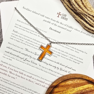 Puede incluir: Un collar plateado con un colgante de cruz naranja con borde plateado. El collar se muestra sobre una tarjeta blanca con texto y un posavasos de madera. El colgante de cruz es el punto focal.