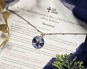 May include: A silver necklace with a round pendant featuring a winter scene with a Christmas tree. The necklace rests on a printed devotional with text about faith and Christmas. Decorative greenery and a blue cloth are in the background.