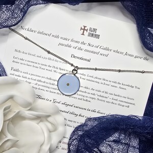 May include: A silver necklace with a pendant containing a mustard seed. The pendant is a light blue circle with a small, round, yellow seed in the center. The necklace is displayed with a devotional text and a white flower.