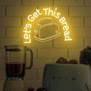 May include: A yellow neon sign with the text "Let's Get This Bread" and a toaster illustration. The sign is hanging above a blue toaster and a blender.