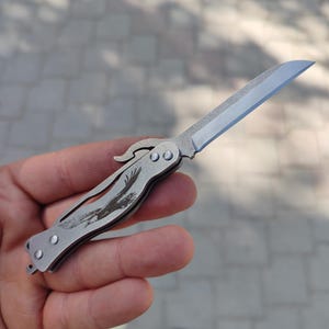 以下が含まれることがあります： 鋭い刃が付いた、小さな銀色の折りたたみナイフ。 ハンドルには、彫刻されたワシのデザインがあります。 ナイフは開いており、刃が表示されています。 ナイフは金属製のように見えます。