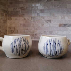 May include: Two white ceramic mugs with a rounded shape and a handle. Each mug features a blue abstract design of vertical lines and dots. The mugs are displayed on a grey countertop against a neutral tile backsplash.