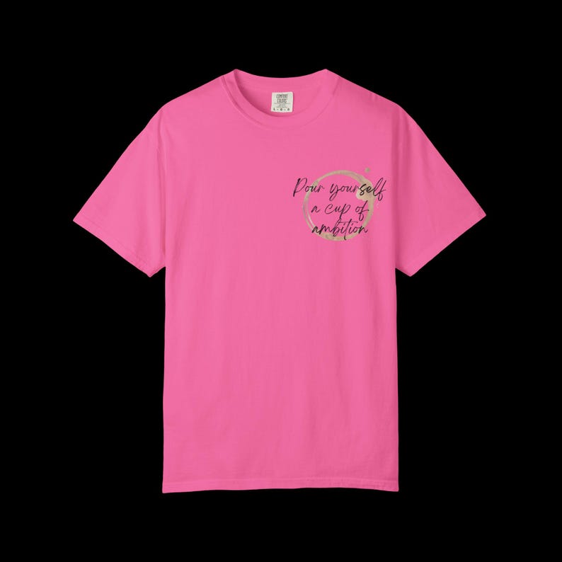 May include: A bright pink short-sleeve t-shirt featuring a circular coffee stain graphic on the left chest area. Inside the stain, the black cursive text reads, Pour yourself a cup of ambition. The shirt has a crew neckline and a standard fit.