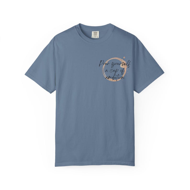 May include: A slate blue short-sleeve t-shirt featuring a graphic on the left chest. The design consists of a light brown circular coffee ring stain with the black cursive text "Pour yourself a cup of ambition" printed inside the circle.