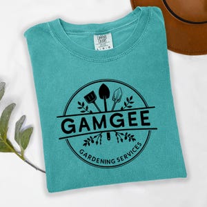 May include: Teal Comfort Colors t-shirt with a black circular graphic. The graphic includes the word "GAMGEE" above "Gardening Services" and illustrations of gardening tools. A brown hat is partially visible in the background.