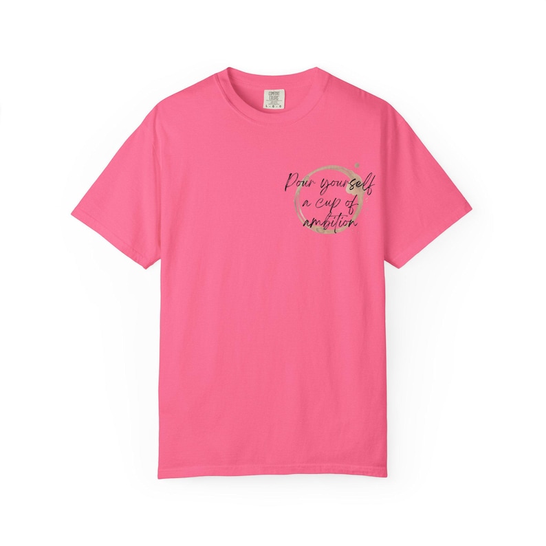 May include: A bright pink short-sleeve t-shirt featuring a circular coffee stain graphic on the upper left chest. Inside the circle, the black cursive text reads, Pour yourself a cup of ambition. The shirt has a crew neckline and a standard fit.