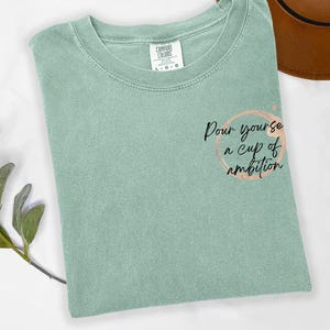 May include: A sage green short-sleeve t-shirt with a crew neckline. The shirt features a graphic on the right side consisting of a light brown circular coffee ring stain and the black cursive text, Pour yourse a cup of ambition.