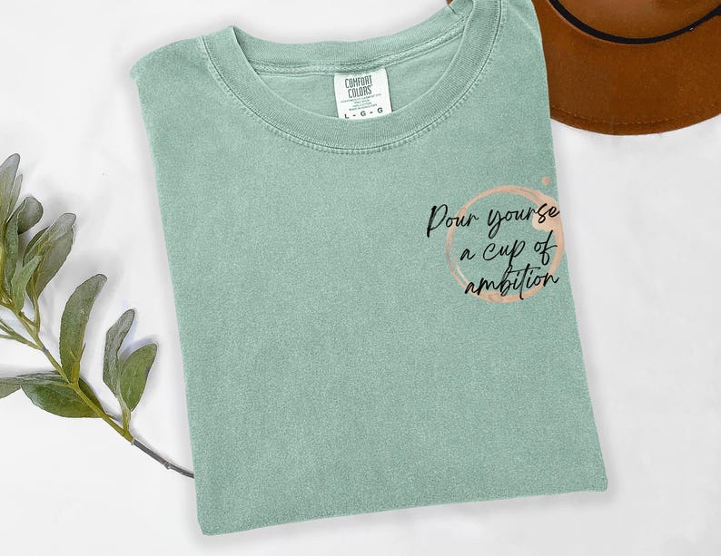 May include: A sage green short-sleeve t-shirt with a crew neckline. The shirt features a graphic on the right side consisting of a light brown circular coffee ring stain and the black cursive text, Pour yourse a cup of ambition.