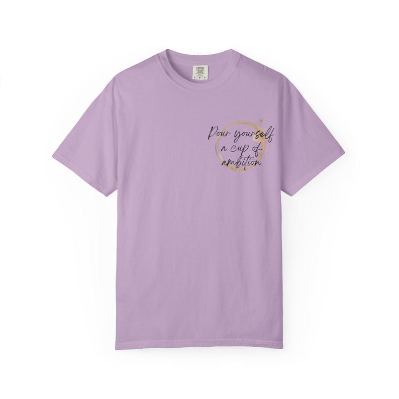 May include: A light purple short-sleeve t-shirt featuring a gold circular coffee ring stain graphic on the left chest. Inside the circle, the black cursive text reads, "Pour yourself a cup of ambition." The shirt has a crew neckline and a standard fit.