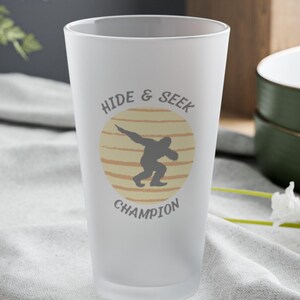 Puede incluir: Vaso de pinta esmerilado con las palabras "HIDE & SEEK CHAMPION" y una silueta de una persona haciendo dab dentro de un círculo amarillo y naranja. El vaso está sobre un paño gris.