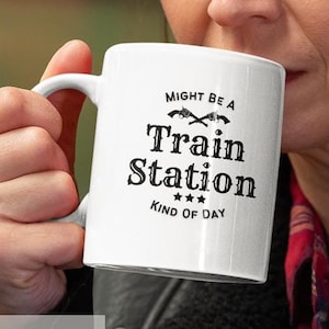 Könnte beinhalten: Eine weiße Keramiktasse mit einem schwarzen Design, auf dem "Might be a Train Station Kind of Day" steht. Die Tasse hat ein schwarzes Kreuzgitterdesign mit zwei Pfeilen, die in entgegengesetzte Richtungen zeigen. Die Tasse wird von einer Person gehalten, die ein rot-schwarz kariertes Hemd und eine schwarze Lederjacke trägt.