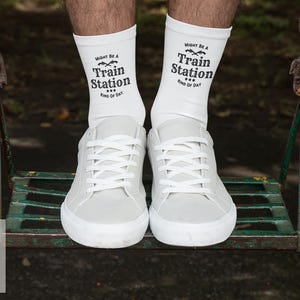 May include: White crew socks with the text "Might Be A Train Station... Kind of Day" are worn with white sneakers. The image shows the lower legs and feet of a person on a swing set.