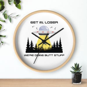 May include: A round wall clock with a light wood frame. The clock face features a cartoon UFO design with the text "GET IN, LOSER" above and "WE'RE DOING BUTT STUFF" below the image.