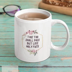 May include: White ceramic coffee mug with a gold hexagonal frame and pink floral design. The text inside the frame reads "This too shall pass but like holy fuck."