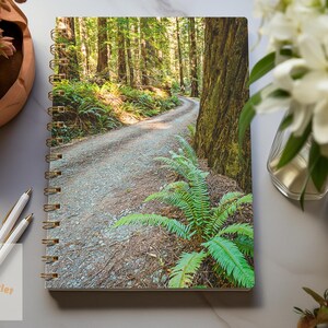 May include: A spiral bound notebook with a photo of a winding dirt road through a lush green forest. The road is lined with ferns and tall trees.