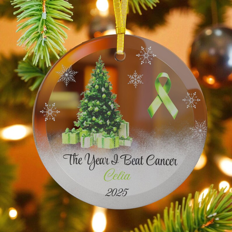 May include: A round glass Christmas ornament with a green ribbon, Christmas tree, and gifts. The text reads "The Year I Beat Cancer Celia 2025". The ornament hangs from a gold ribbon.