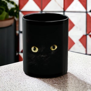 May include: Black ceramic mug with a black cat peeking out from behind the rim. The cat has bright yellow eyes.