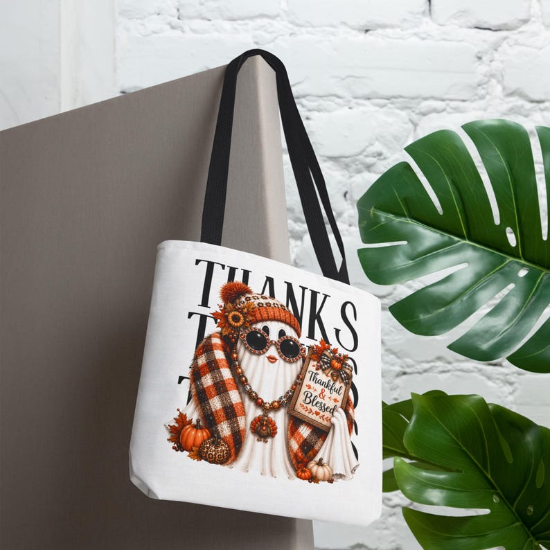 May include: A white tote bag with black handles featuring a graphic of a ghost wearing a plaid scarf, a beanie, and sunglasses. The ghost holds a sign that reads Thankful & Blessed, surrounded by pumpkins, sunflowers, and autumn leaves.