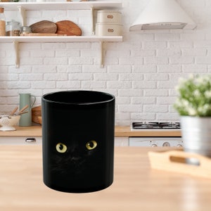 May include: Black ceramic mug with a black cat peeking out from behind the mug. The cat has bright yellow eyes.