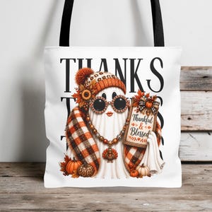 May include: A white tote bag with black straps featuring a graphic of a ghost wearing a plaid scarf, a beanie, and round sunglasses. The ghost holds a sign that reads Thankful & Blessed, surrounded by pumpkins, sunflowers, and autumn leaves.