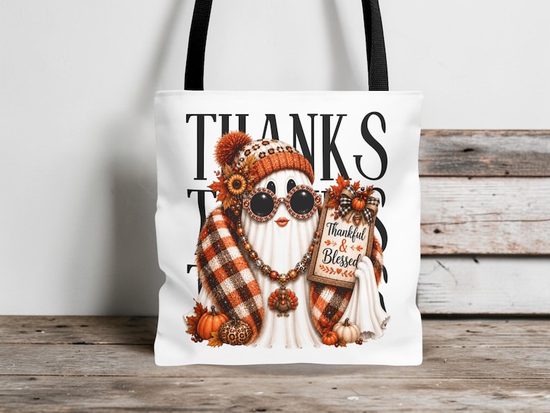 May include: A white tote bag with black straps featuring a graphic of a ghost wearing a plaid scarf, a beanie, and round sunglasses. The ghost holds a sign that reads Thankful & Blessed, surrounded by pumpkins, sunflowers, and autumn leaves.