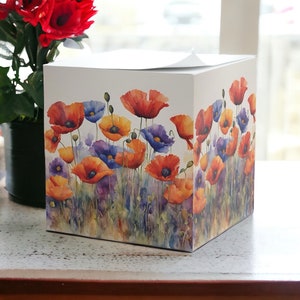 May include: A square tissue box featuring a watercolor floral design of red, orange, yellow, and purple poppies. The flowers are set against a white background, with green foliage at the base. The box is sitting on a white surface.