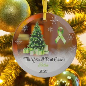 May include: A clear glass Christmas ornament with a green Christmas tree and wrapped gifts. The ornament has a green ribbon and the text "The Year I Beat Cancer Celia 2025". Snowflakes are also featured.