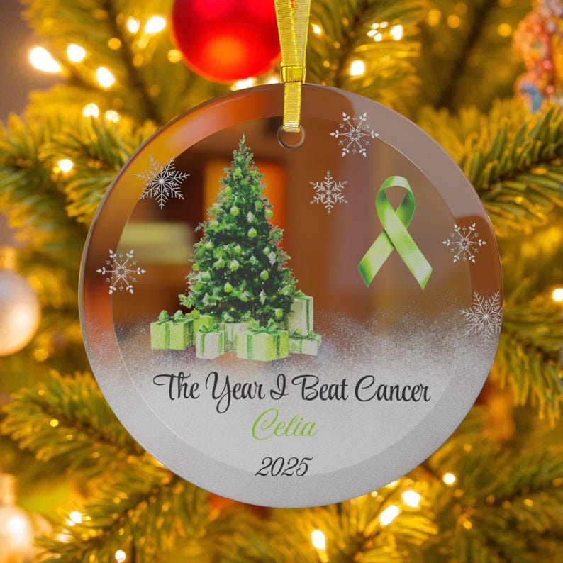 May include: A clear, round Christmas ornament with a green Christmas tree and wrapped gifts. The ornament has a green ribbon and snowflakes. Text reads "The Year I Beat Cancer Celia 2025".