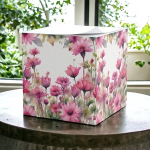 May include: A square, white notepad with a pink floral design. The design features watercolor pink flowers with green leaves and stems on a white background.