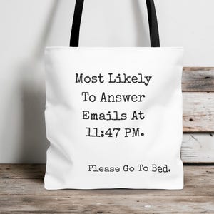 Op de afbeelding: Witte draagtas met zwarte handvatten. De tas heeft de tekst "Most Likely To Answer Emails At 11:47 PM. Please Go To Bed." in het zwart, gecentreerd op de voorkant. De tas staat op een houten oppervlak.