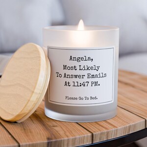 May include: A lit, frosted glass candle with a wooden lid, resting on a wooden table. The candle has a white label with black text that reads: "Angela, Most Likely To Answer Emails At 11:47 PM. Please Go To Bed."