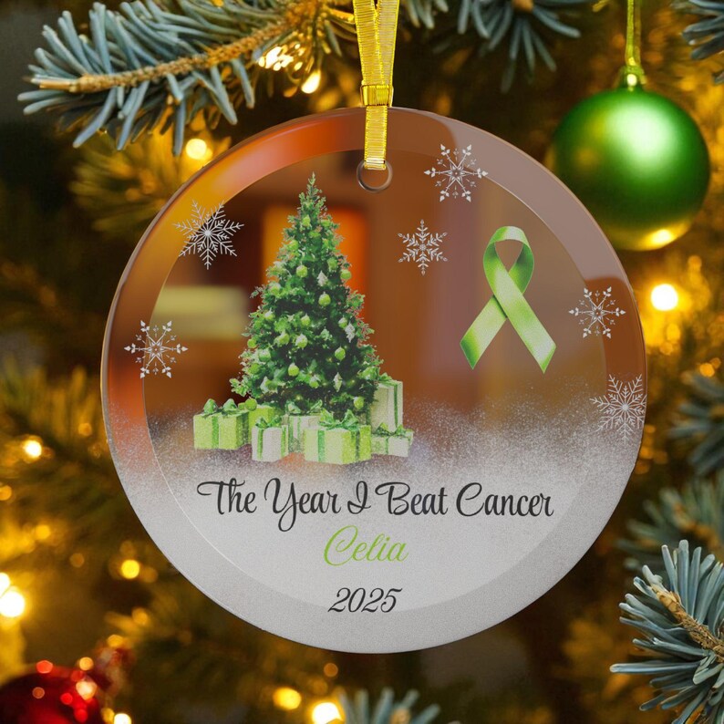 May include: A clear glass Christmas ornament with a gold ribbon. The ornament features a green Christmas tree, gifts, snowflakes, and a green ribbon. Text reads "The Year I Beat Cancer Celia 2025."