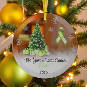 May include: A clear, round Christmas ornament with a gold ribbon. The ornament features a green Christmas tree, gifts, snowflakes, and a green ribbon. Text reads "The Year I Beat Cancer Celia 2025."