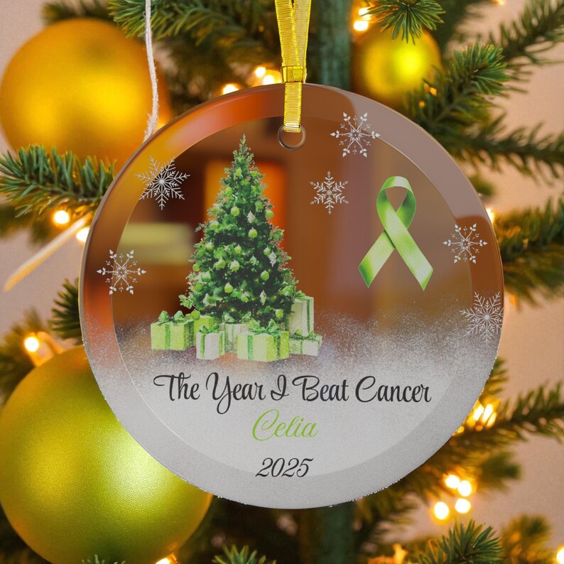 May include: A clear, round Christmas ornament with a gold ribbon. The ornament features a green Christmas tree, gifts, snowflakes, and a green ribbon. Text reads "The Year I Beat Cancer Celia 2025."