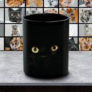 May include: Black ceramic mug with a black cat peeking out from behind the mug, with only its yellow eyes visible.