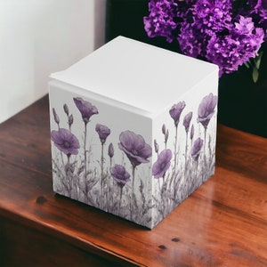Puede incluir: Una caja cuadrada blanca con un diseño floral. La caja está decorada con un patrón de flores silvestres moradas sobre un fondo blanco.