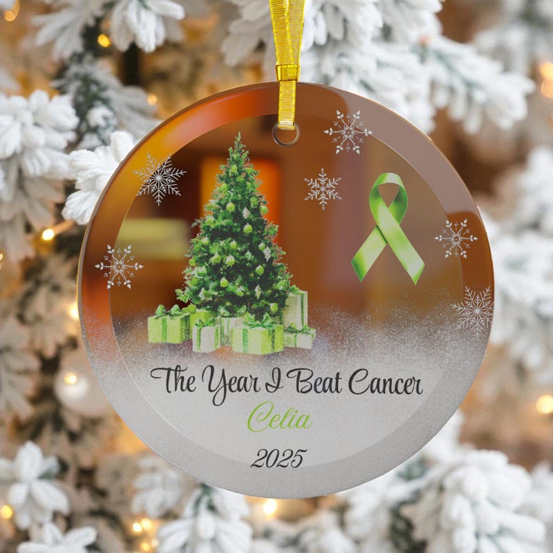 May include: Round glass Christmas ornament with a green Christmas tree, gifts, and snowflakes. Features a green ribbon and the text "The Year I Beat Cancer Celia 2025". Includes a gold ribbon for hanging.