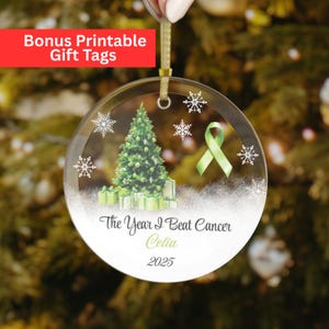 May include: A clear glass Christmas ornament with a green ribbon, Christmas tree, and gift boxes. The text reads "The Year I Beat Cancer Celia 2025". The ornament is hanging from a gold ribbon.