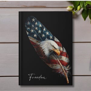 May include: Black journal featuring a detailed illustration of an American flag incorporated into a feather, with an eagle's head. The word "Freedom" is written in cursive. A blue pen and pink flower are on a wooden surface.