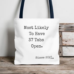Op de afbeelding: Witte draagtas met marineblauwe handvatten. De tas heeft de tekst "Most Likely To Have 37 Tabs Open. Since 2021." in het zwart, gecentreerd op de voorkant. De tas staat op een houten oppervlak.