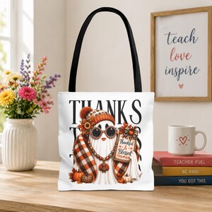 May include: A white tote bag with black handles featuring a graphic of a ghost wearing a plaid scarf, a beanie, and round sunglasses. The ghost holds a sign that reads Thankful & Blessed. The design includes pumpkins and autumn leaves.