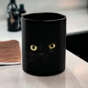May include: Black ceramic coffee mug with a black cat peeking out from the darkness, its yellow eyes staring directly at the viewer.