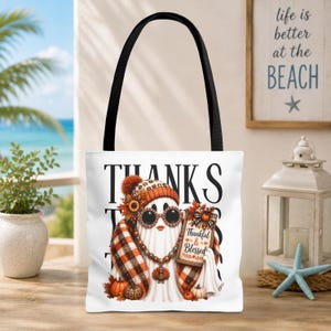 May include: A white tote bag with black handles featuring a graphic of a ghost wearing a plaid scarf, a beanie, and sunglasses. The ghost holds a sign that reads Thankful & Blessed, with the word THANKS printed in large black letters behind it.