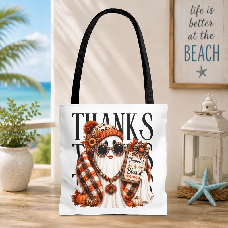 May include: A white tote bag with black handles featuring a graphic of a ghost wearing a plaid scarf, a beanie, and sunglasses. The ghost holds a sign that reads Thankful & Blessed, with the word THANKS printed in large black letters behind it.