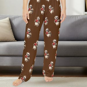 May include: Brown pyjama trousers featuring an all-over repeating pattern of illustrated sloths. Each sloth is depicted in a standing position, holding a small, bright red heart against its chest. The trousers have a relaxed, straight-leg fit.