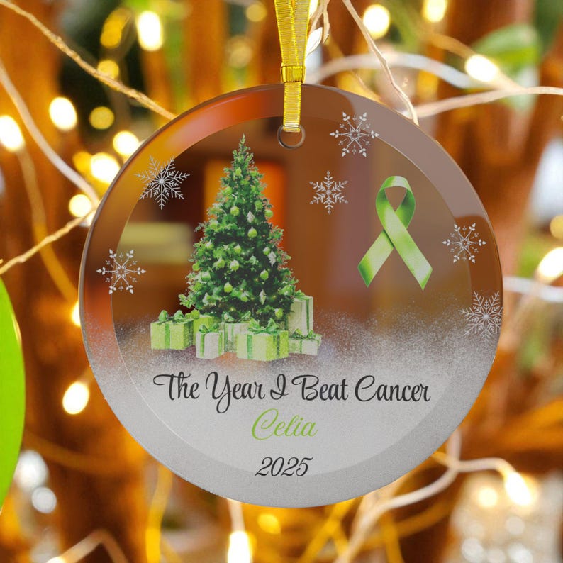 May include: A round glass Christmas ornament with a gold ribbon. The ornament features a green Christmas tree, gifts, snowflakes, and a green ribbon. Text reads "The Year I Beat Cancer Celia 2025."